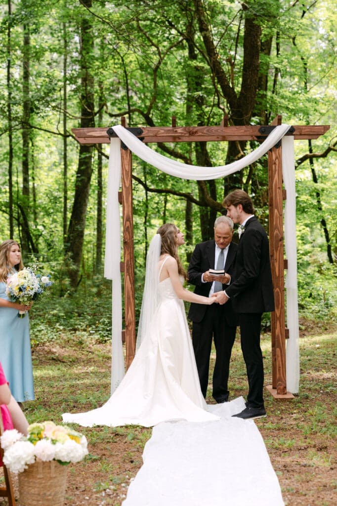 bride and groom exchanging vows with the officiant beneath the wooden arbor in the pines