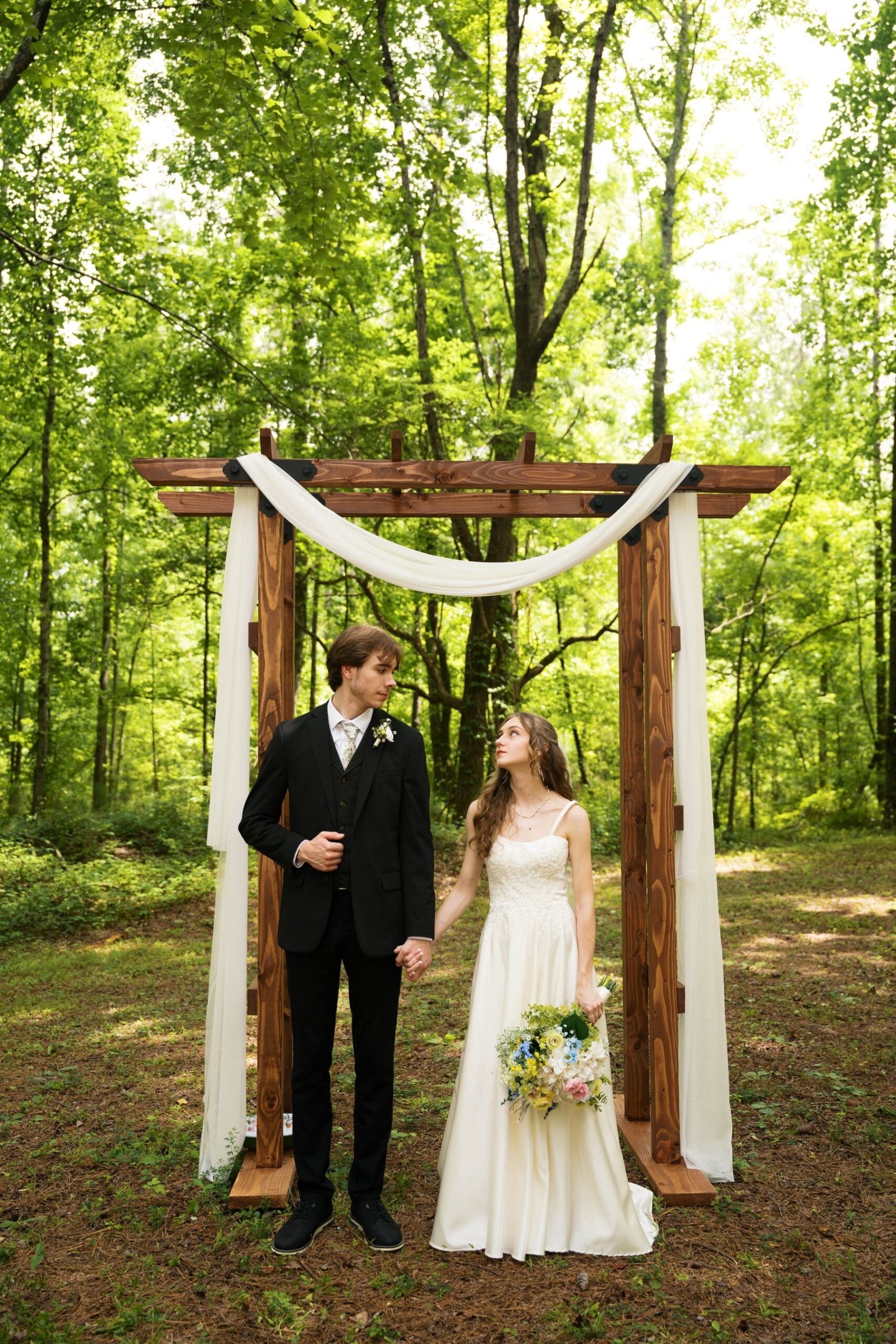 bride and groom beneath a wooden arbor draped in white fabric at their forest wedding